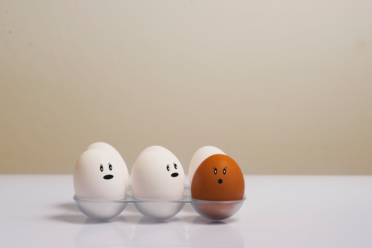 Cartoon of a few white eggs in an egg carton being hostile to a brown egg because it looks different.